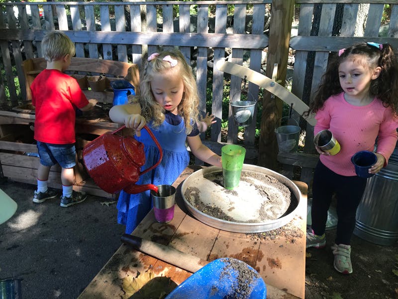 Child pouring water from a watering can into a cup, with other children nearby