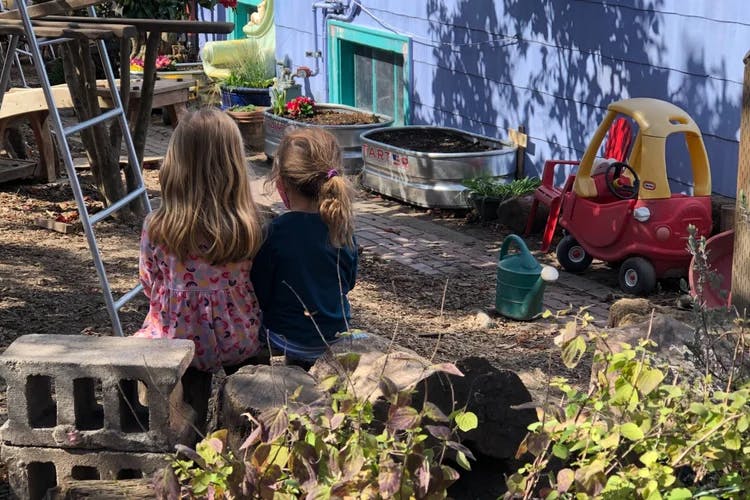 Two children sitting on logs in a garden, with planters and a toy car nearby