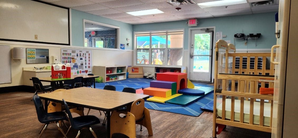 Classroom with child-sized tables and chairs, play mats, and a crib