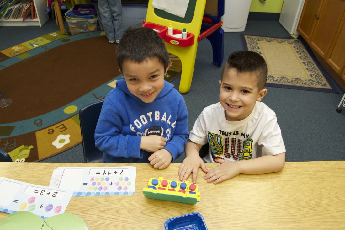 Two children sitting at a table with a toy and papers in a classroom