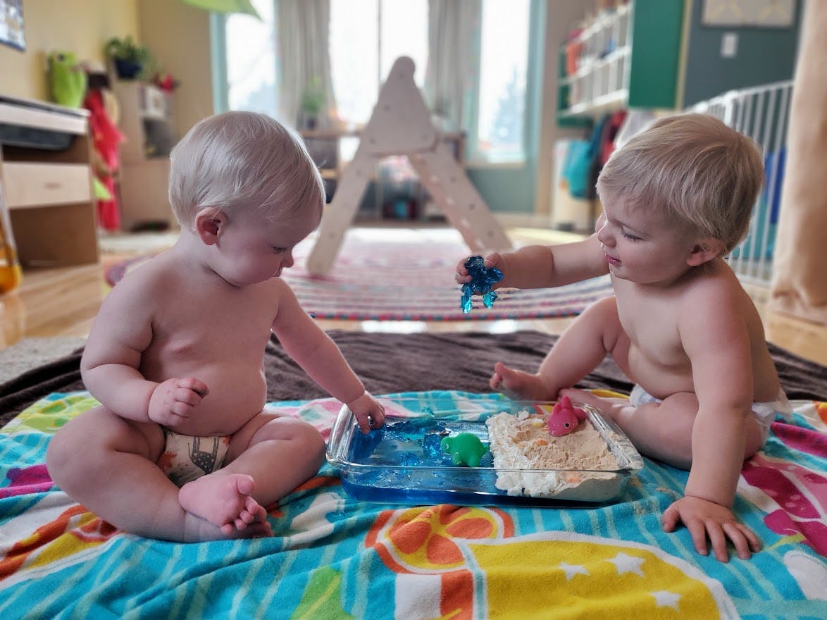 Two babies playing with toys in a sensory bin