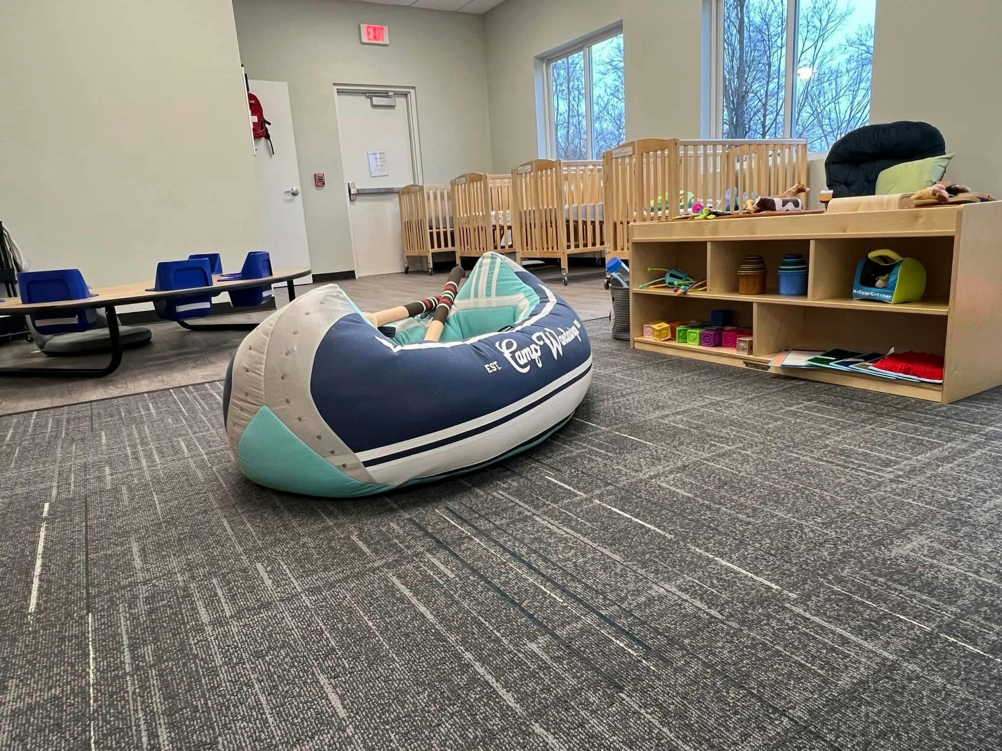 A playroom with cribs, small tables, a storage unit with toys, and a boat-shaped beanbag