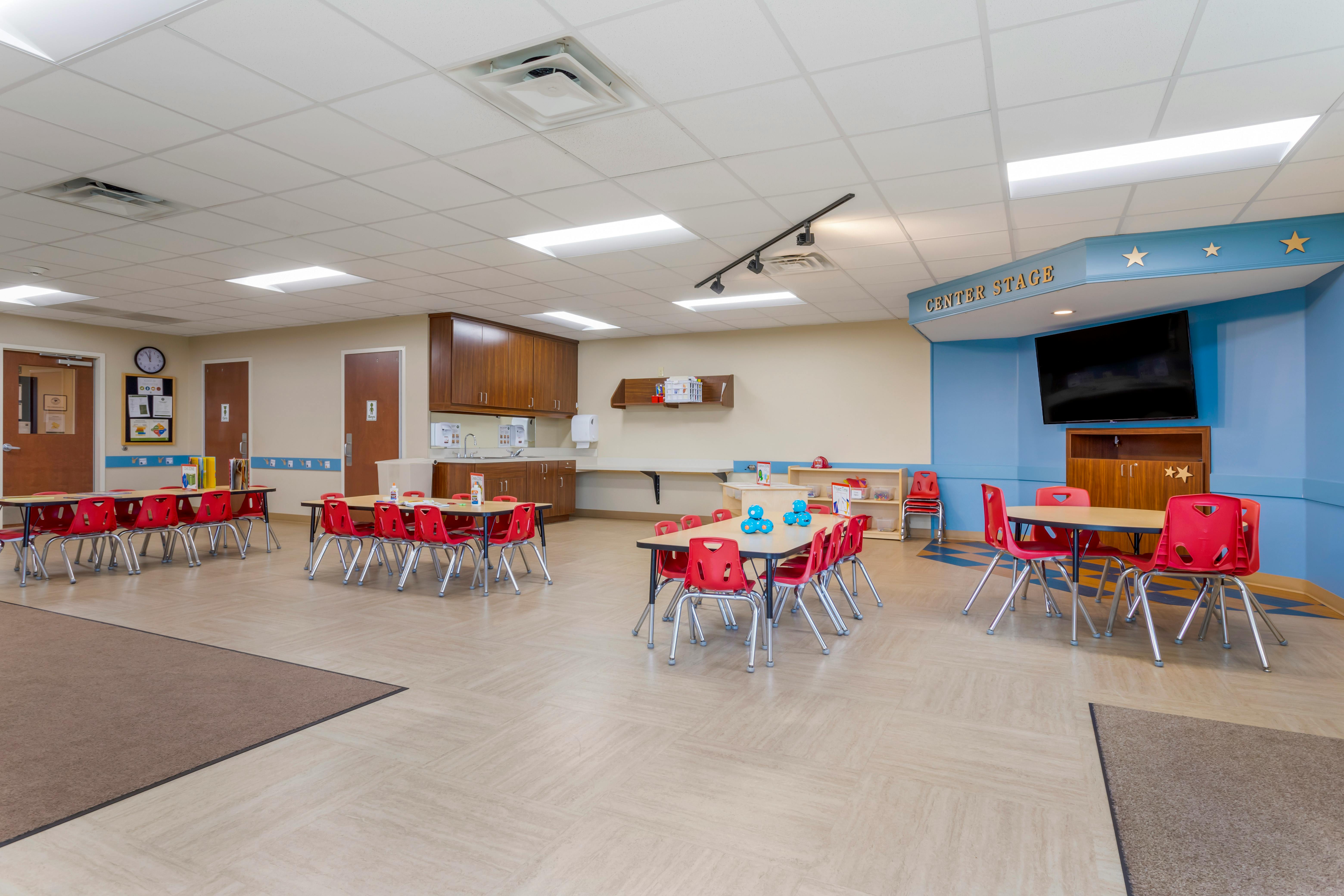Classroom with multiple tables and red chairs, a small stage area, and wall-mounted cabinets