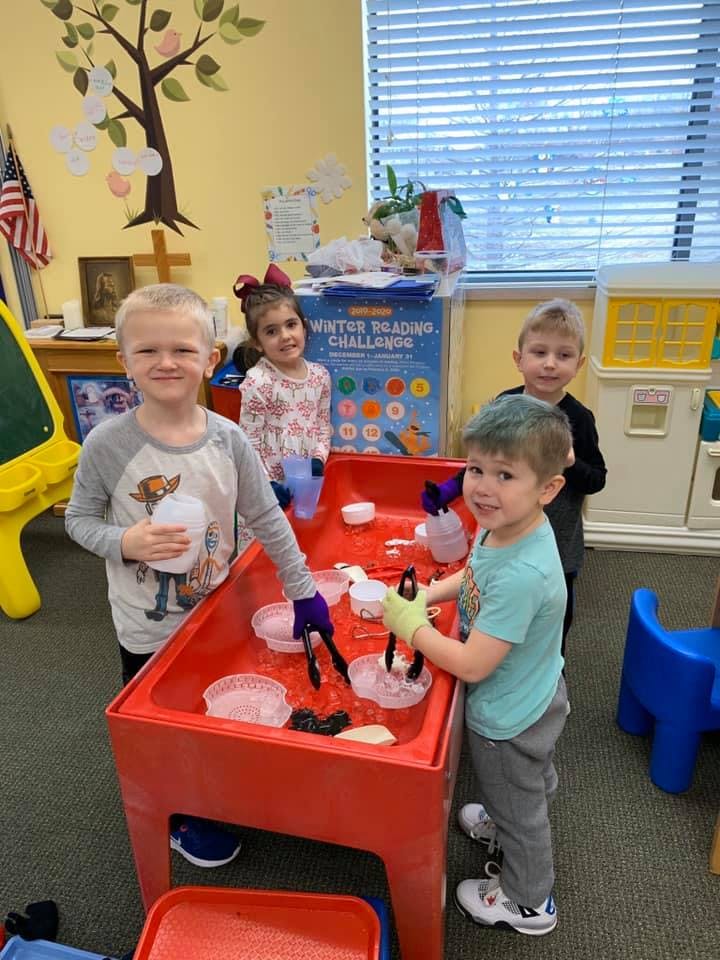 Four children playing at a red sensory table with ice and tongs