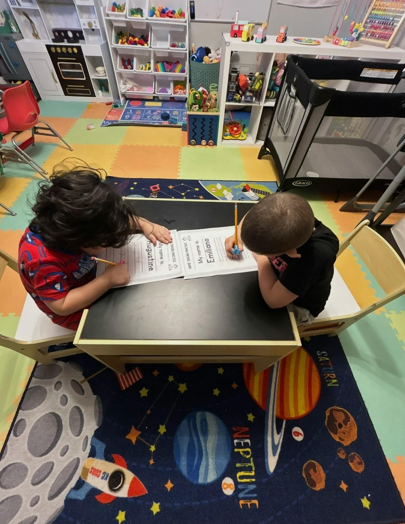 Two children seated at a small table, practicing name tracing in a classroom