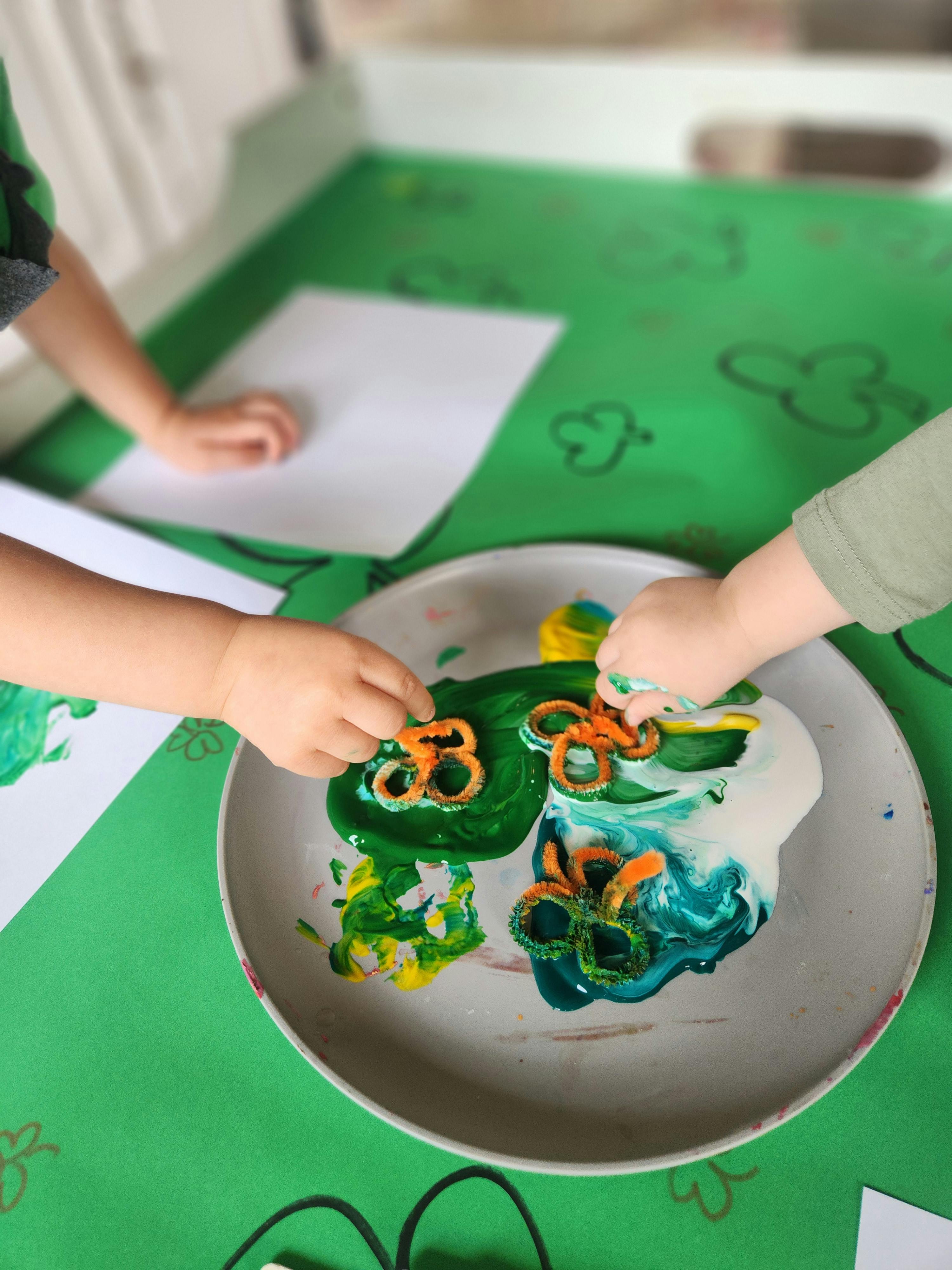 Two children's hands playing with paint and pretzels in a tray