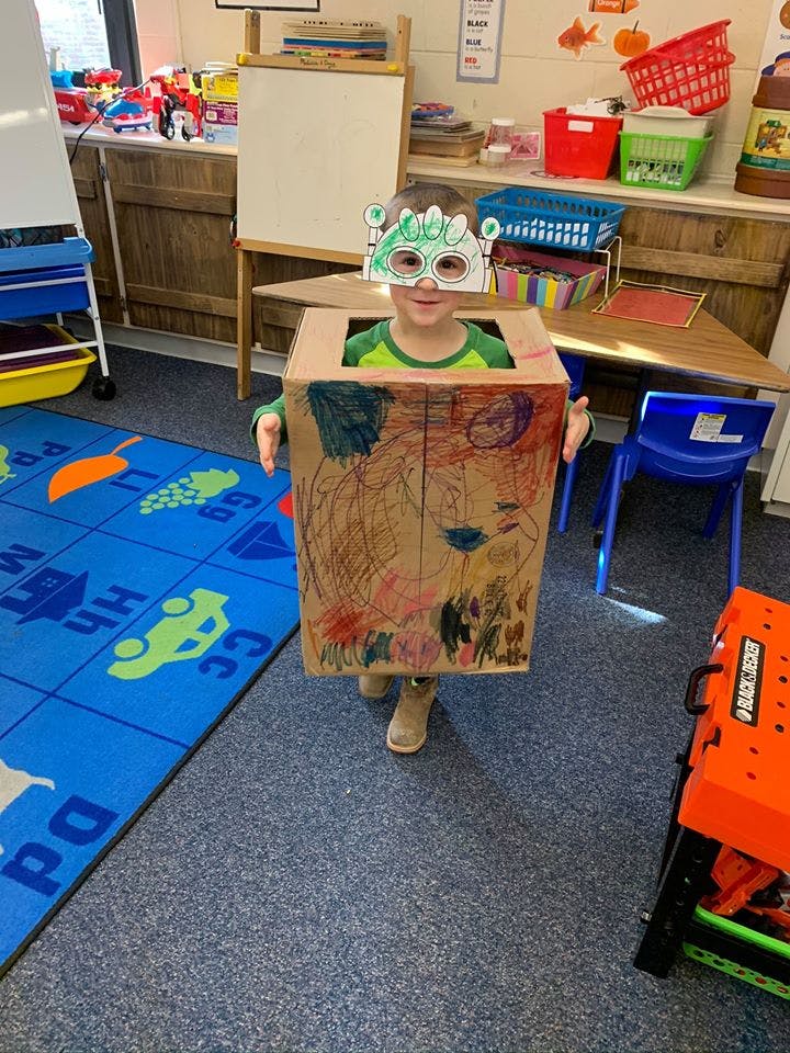 Child wearing a decorated cardboard box and a paper mask in a classroom