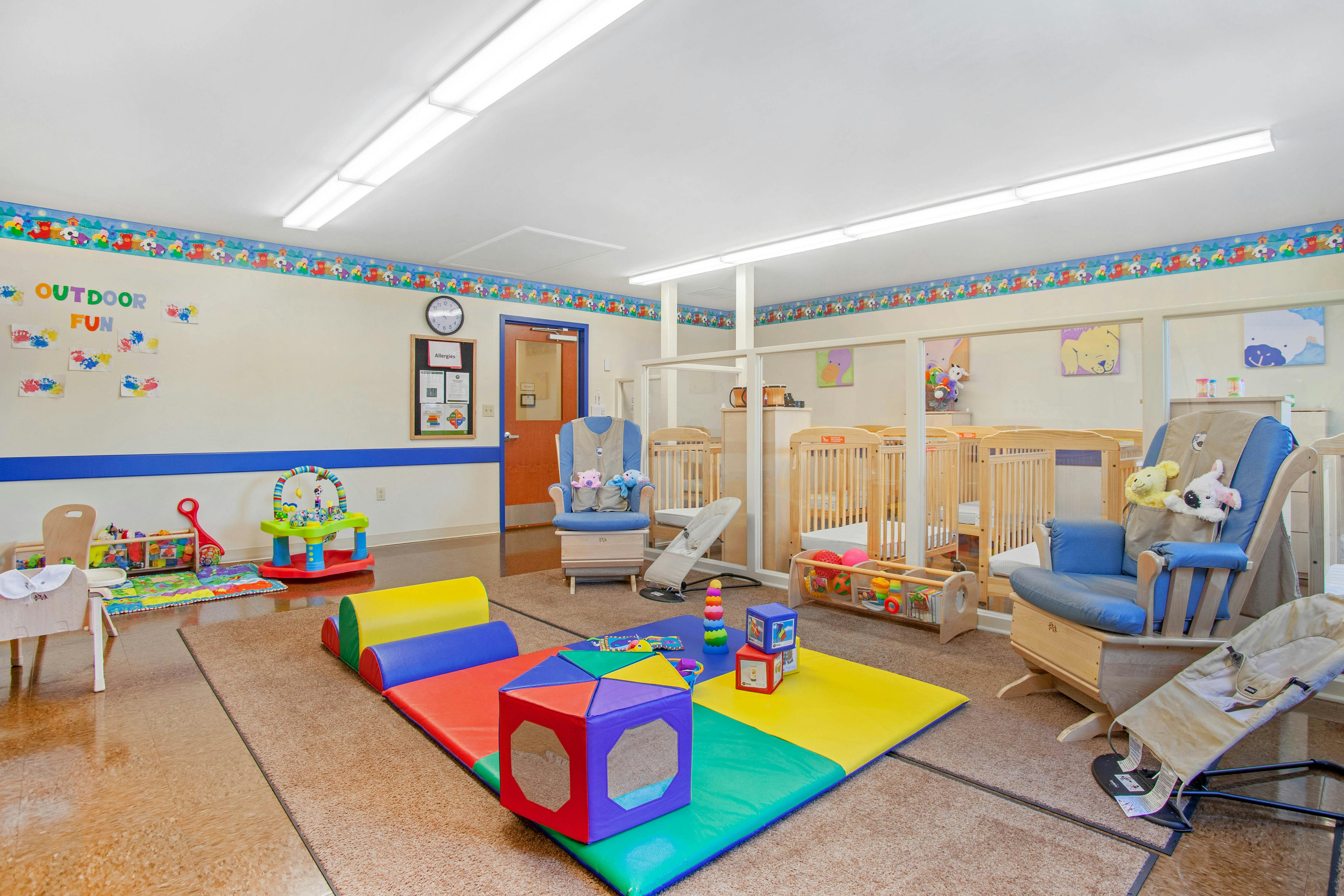A colorful childcare classroom with cribs, playmats, and rocking chairs