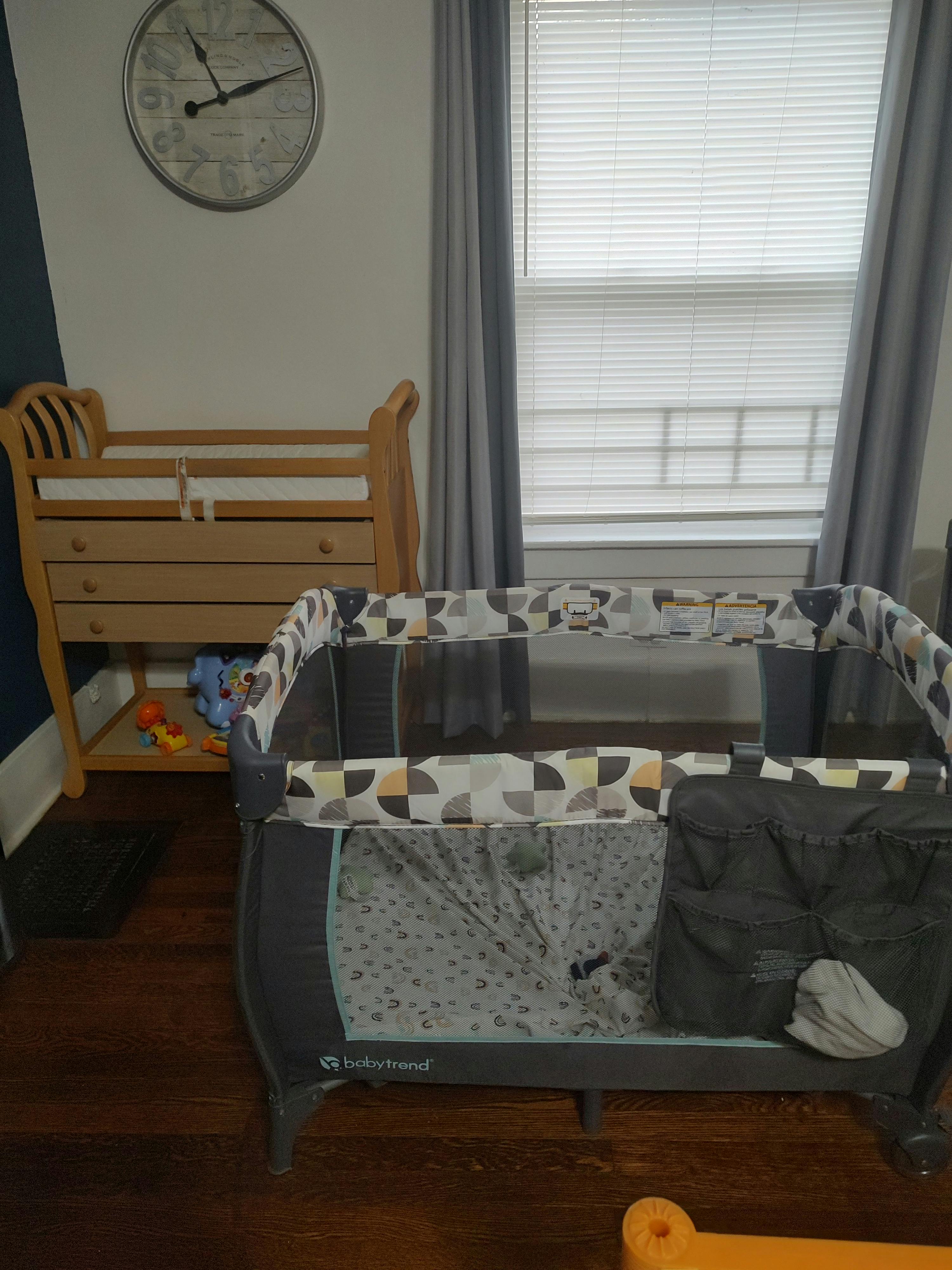 A wooden changing table next to a playard in a room with a window