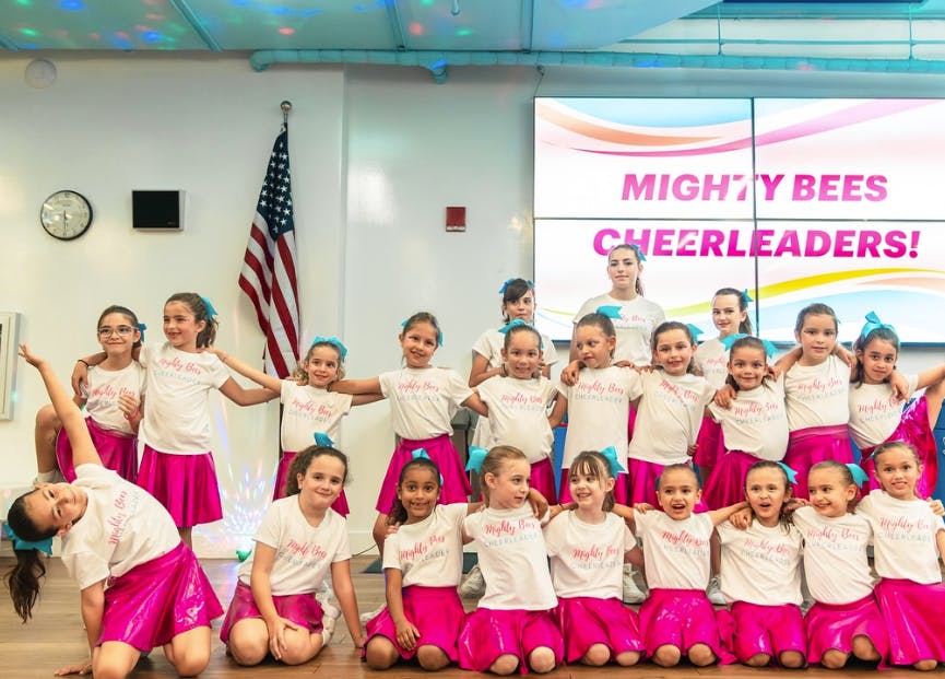 Group of children in white shirts and pink skirts posing, with a screen displaying "MIGHTY BEES CHEERLEADERS!"