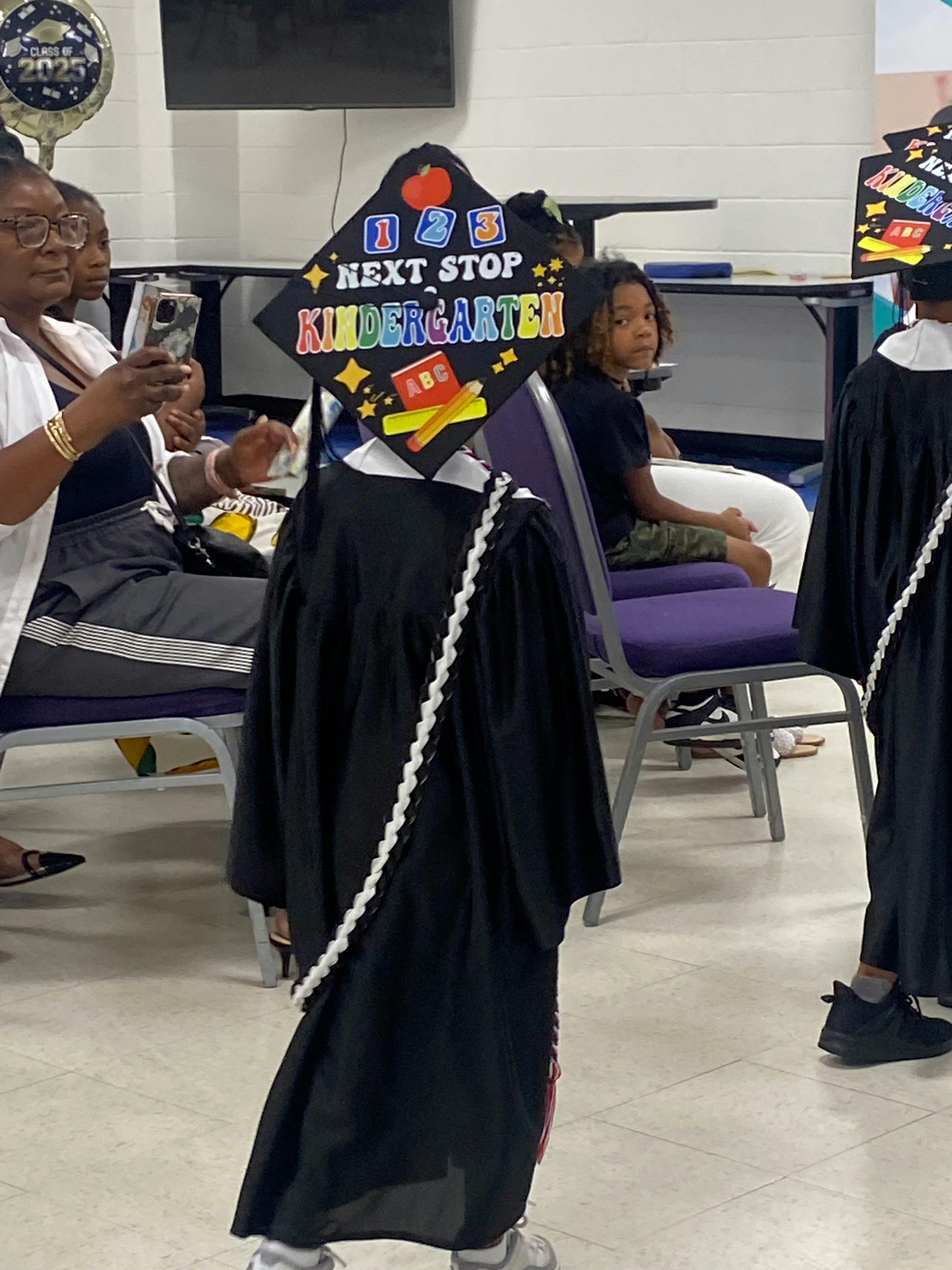 Child wearing graduation regalia and a Next Stop Kindergarten cap