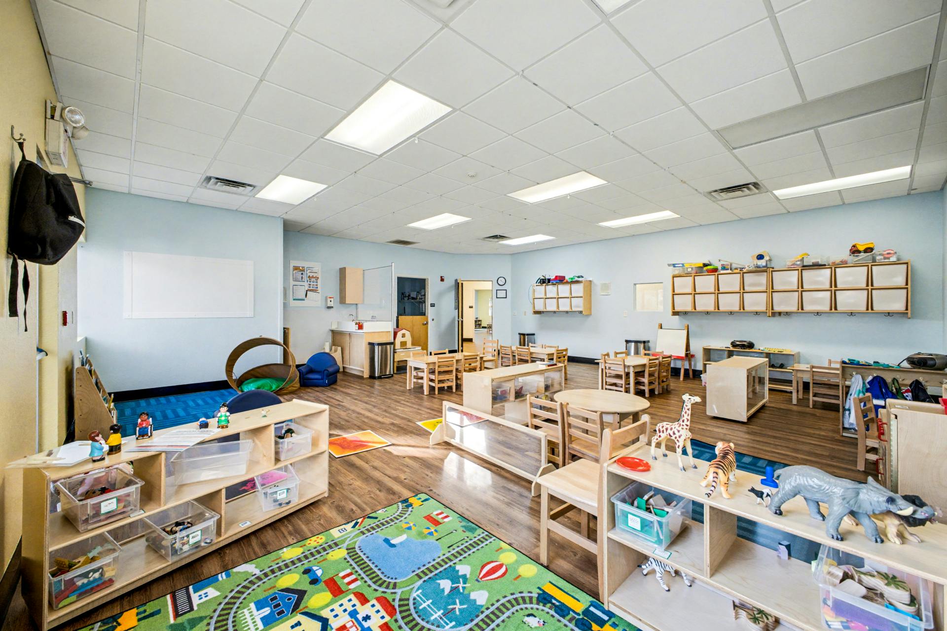 Classroom with small tables and chairs, shelves with toys, and a colorful rug