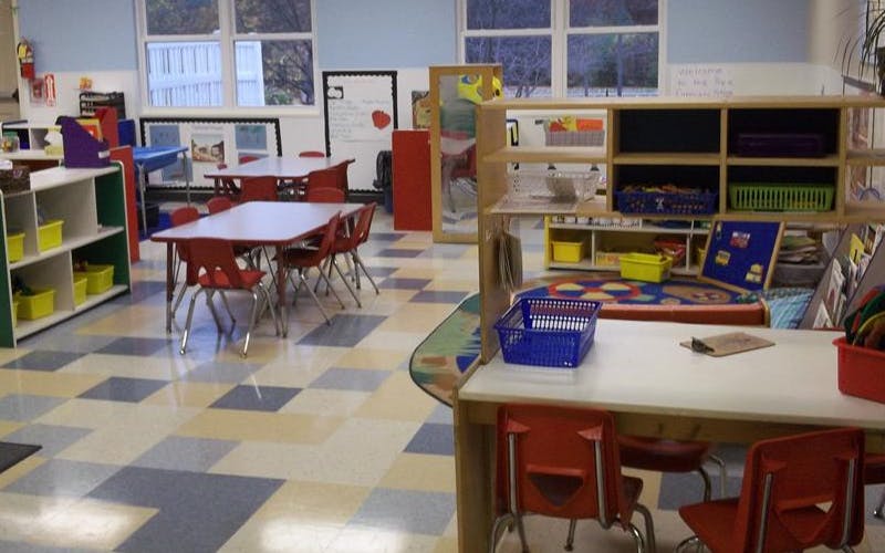 Empty classroom with small red tables and chairs, shelving units with bins