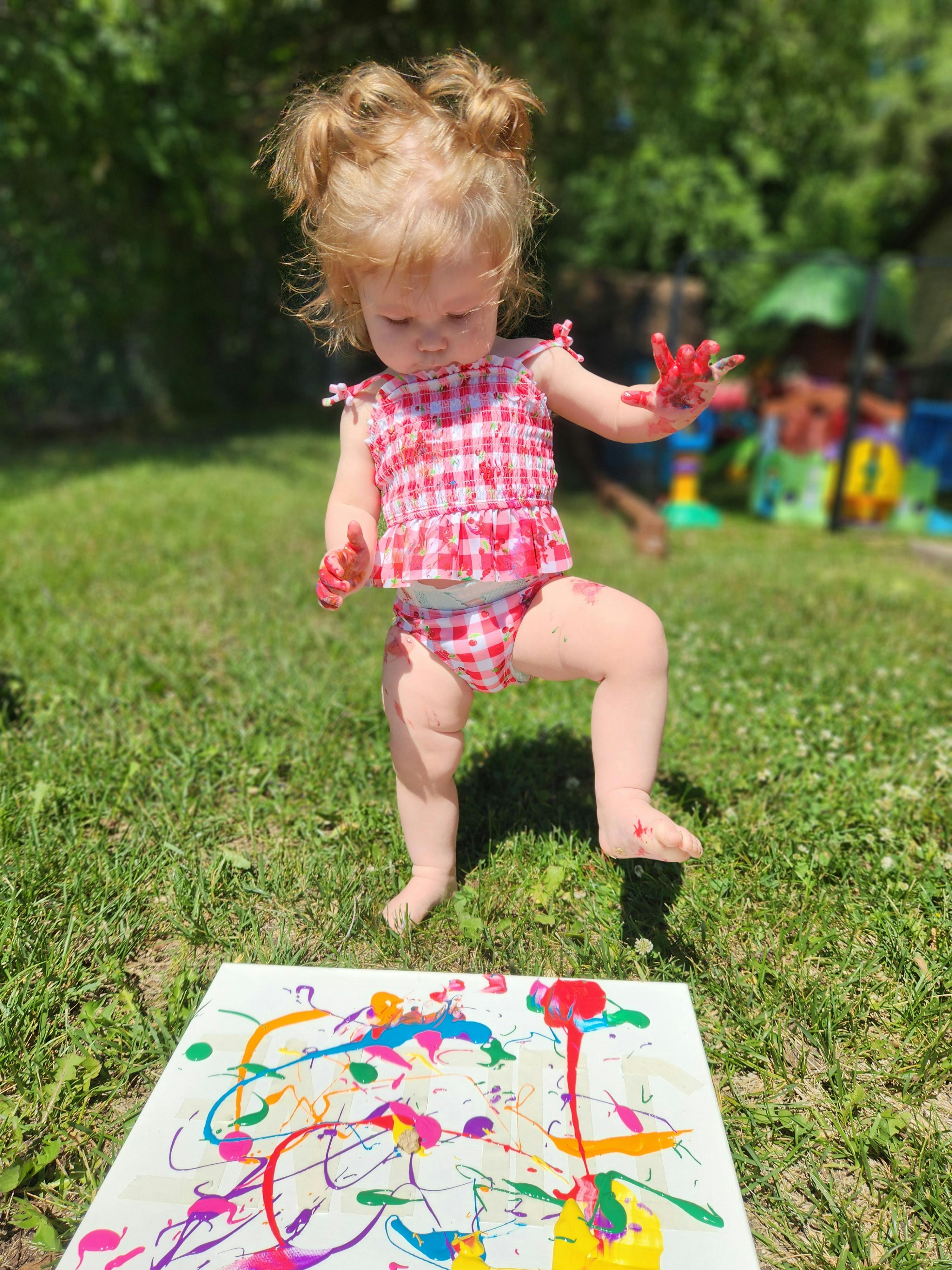 Child with paint on hands standing on grass next to a canvas