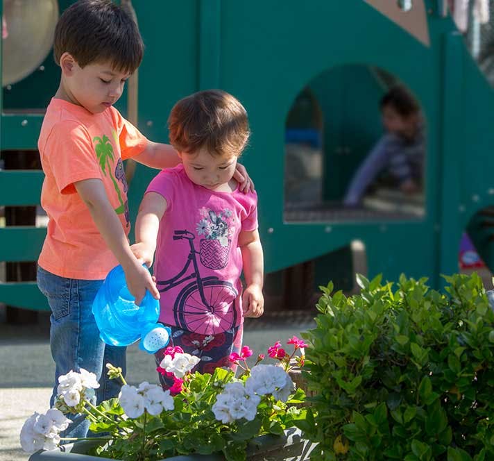Child watering flowers with a small watering can, another child nearby
