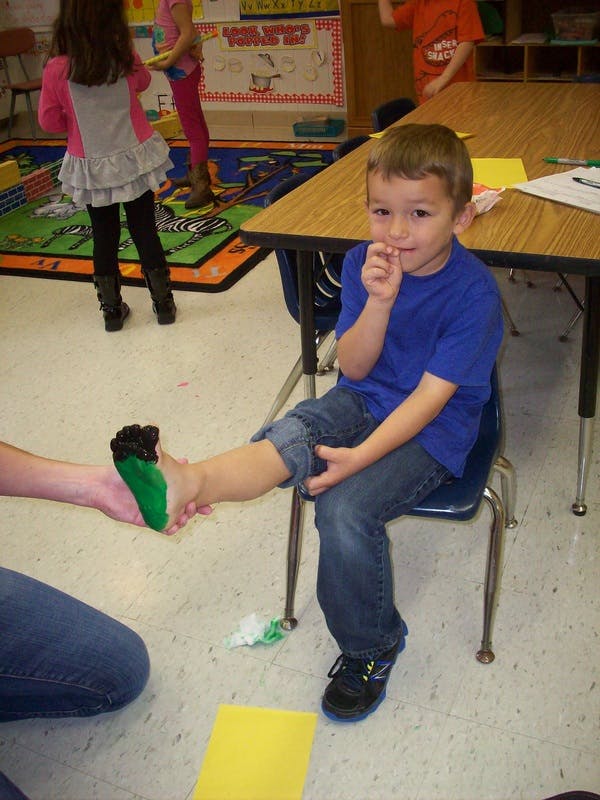 Child sitting in a chair with a person holding their foot covered in green paint
