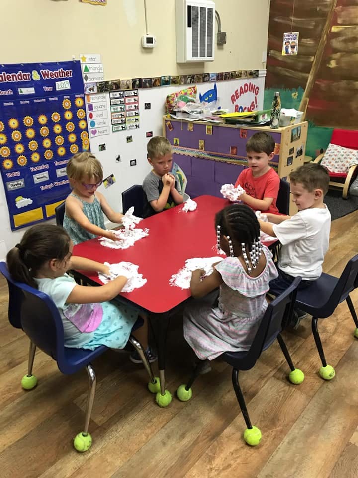 Five children playing with white foam at a red table in a classroom
