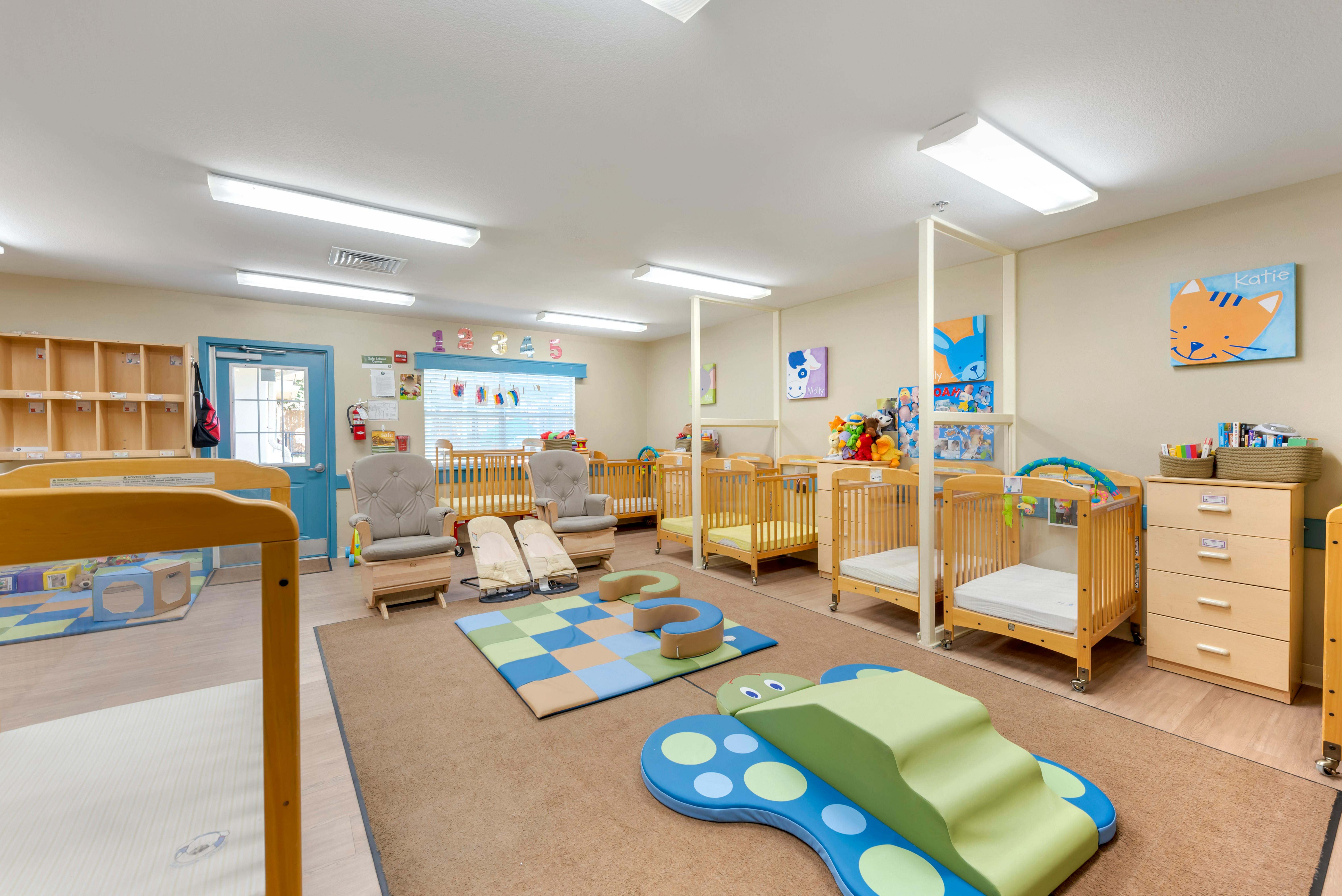 A brightly lit nursery room with multiple cribs, two rocking chairs, and play mats
