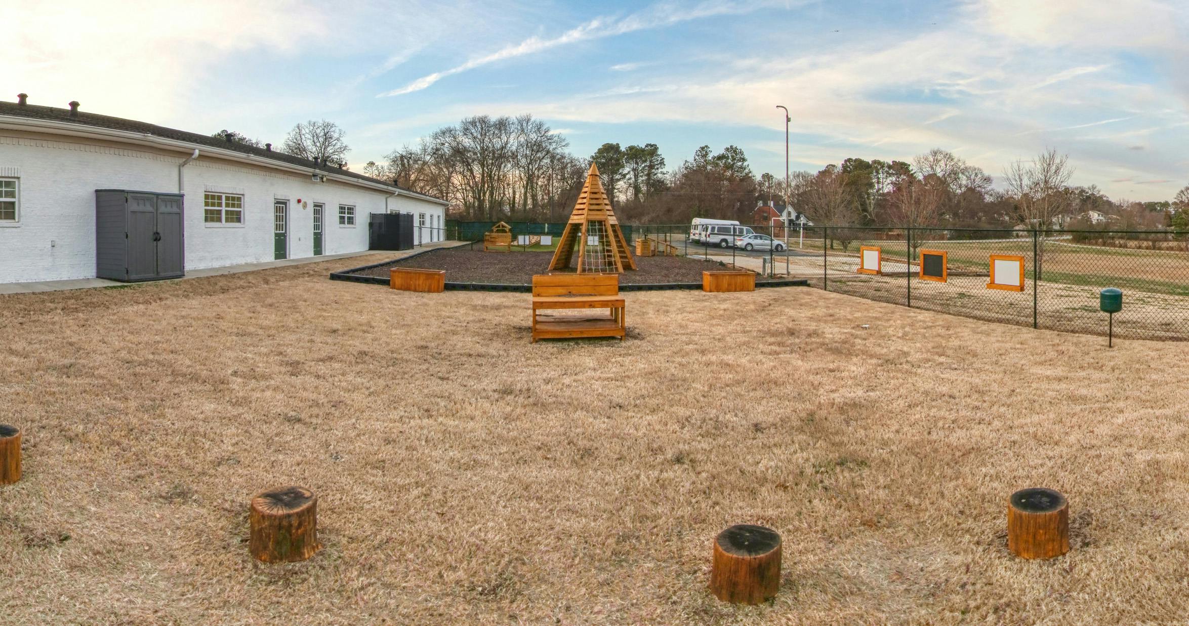 Outdoor play area with a wooden climbing structure, log seats, and a building