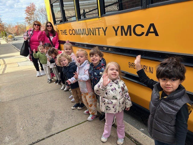 Two people and a line of children standing next to a yellow bus with "YMCA" on it