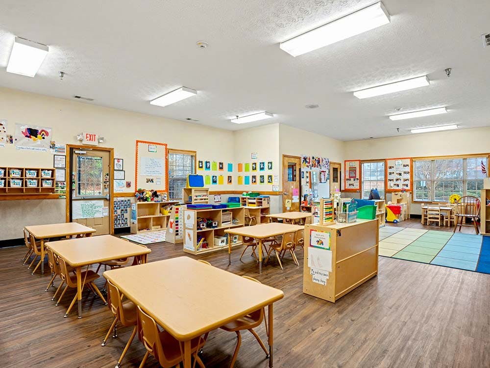 Classroom with small tables and chairs, shelves with toys, and an exit sign above a door