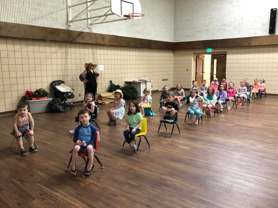 Children sitting on small chairs in a large room with a person standing