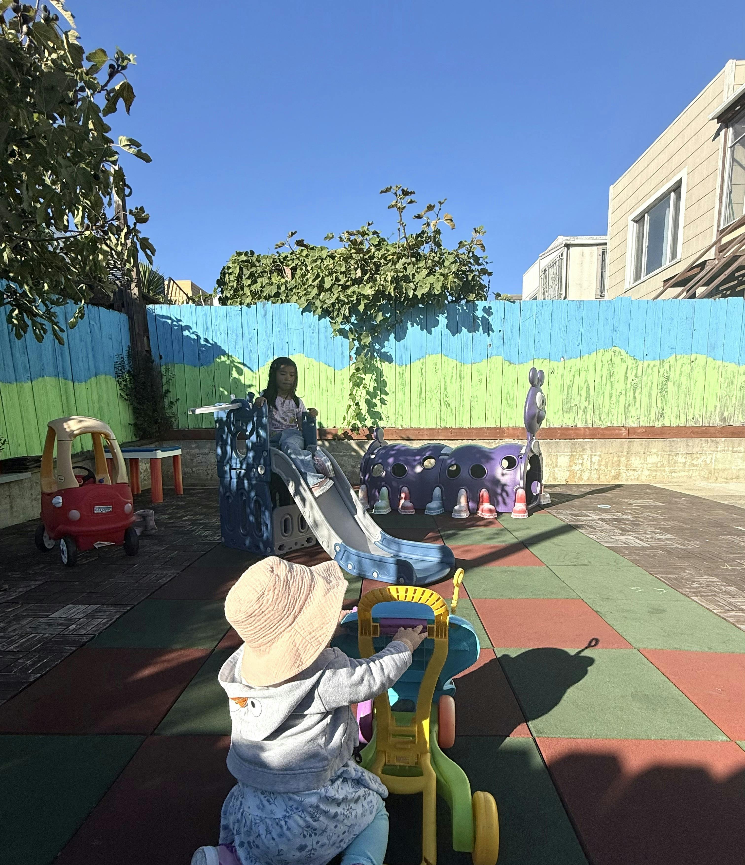 Child playing on a slide while another child pushes a toy walker in a backyard