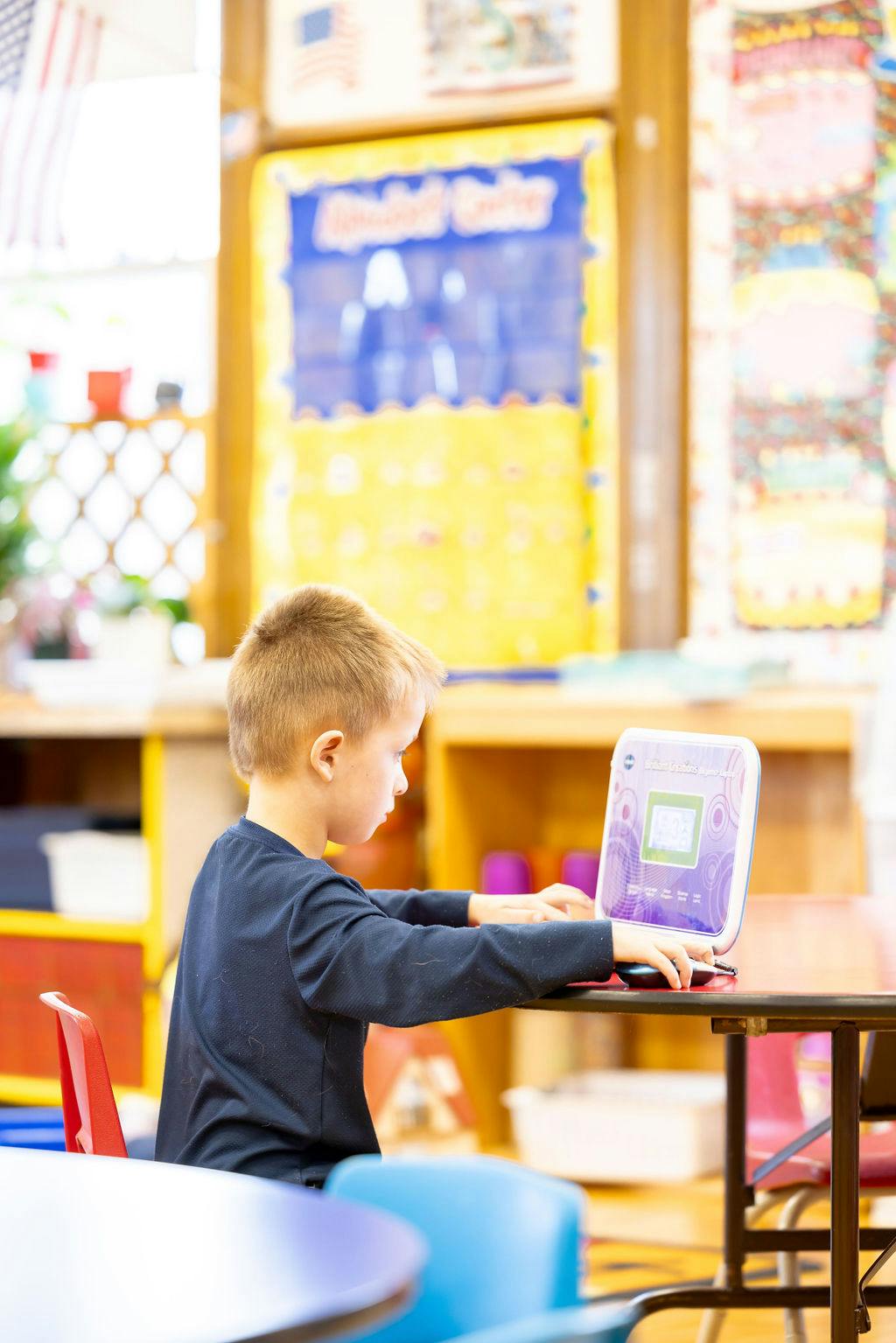 Child operating a small laptop at a table in a classroom