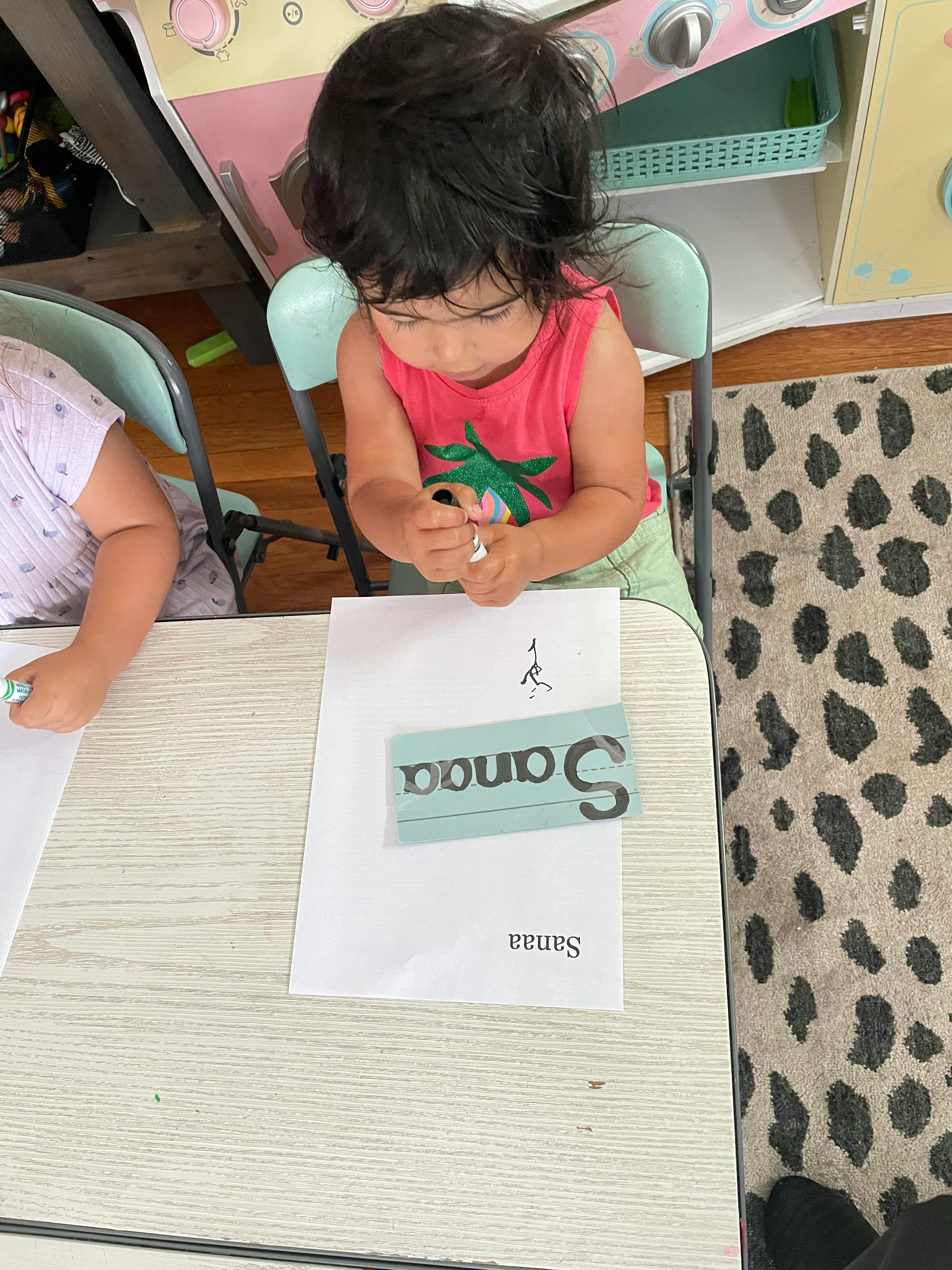 Child sitting at a table, looking down at a paper with the word Sanaa