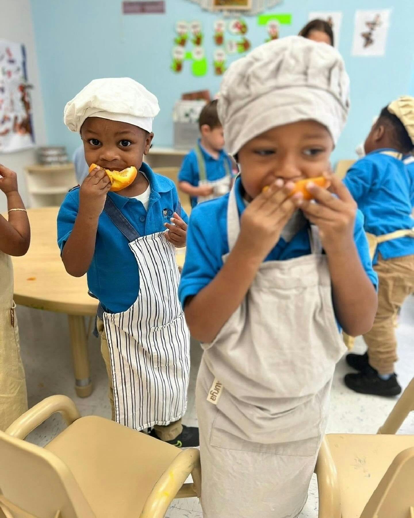 Two children wearing chef hats and aprons eating orange slices