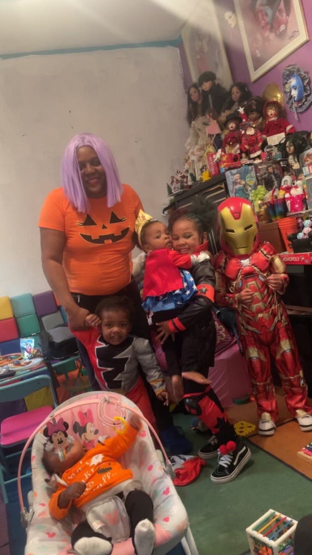 Person in pumpkin shirt with three children, one in Iron Man costume, one in baby bouncer
