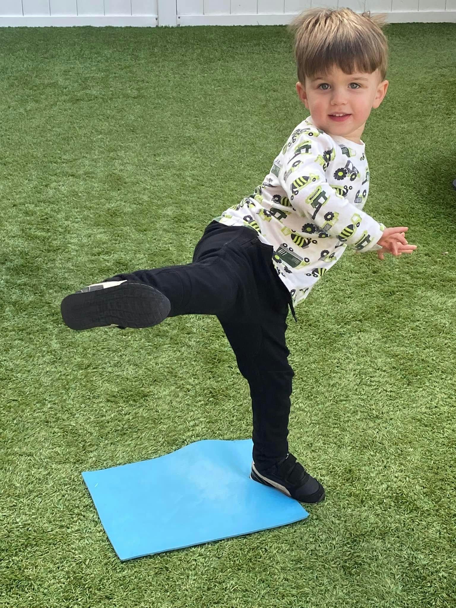 Child balancing on one leg on a blue mat on artificial grass