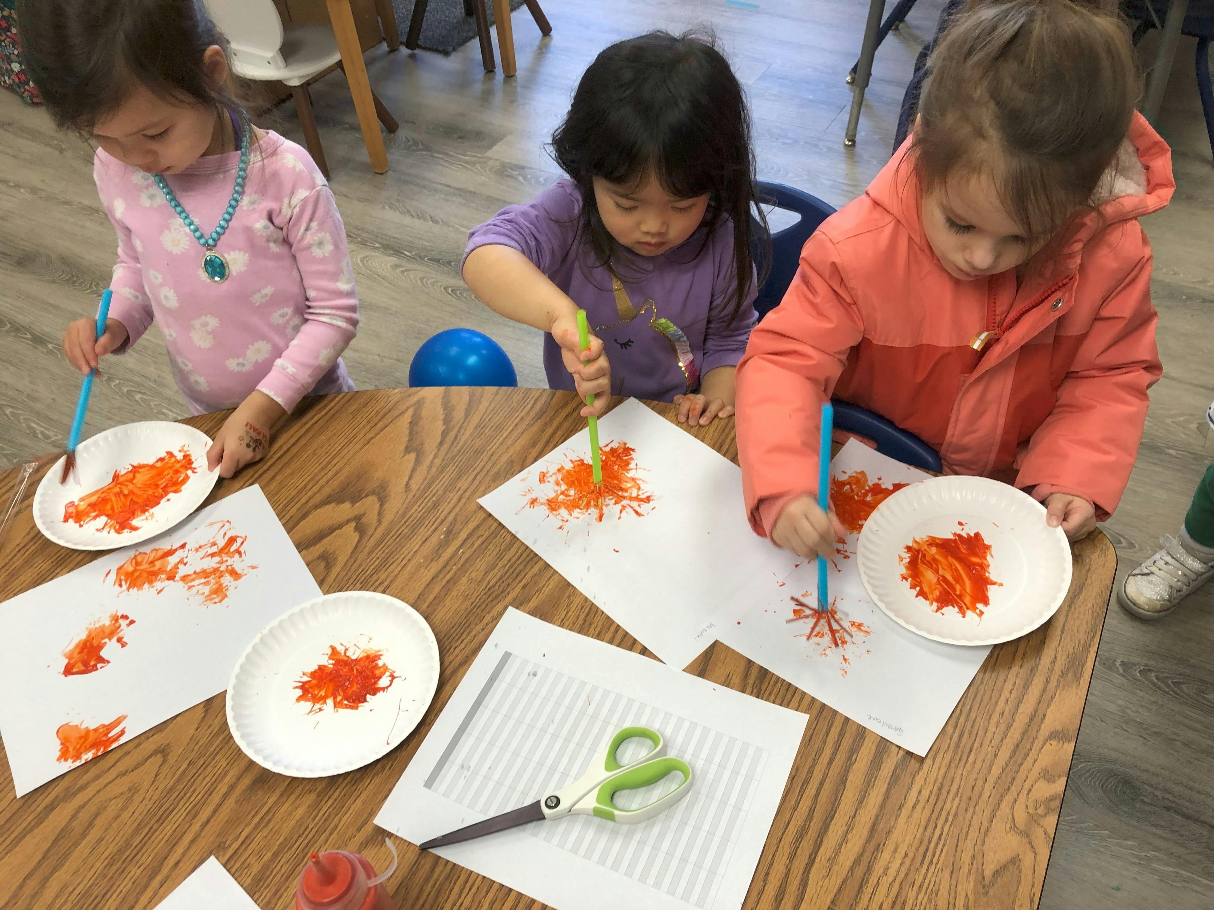 Three children painting with orange paint on paper
