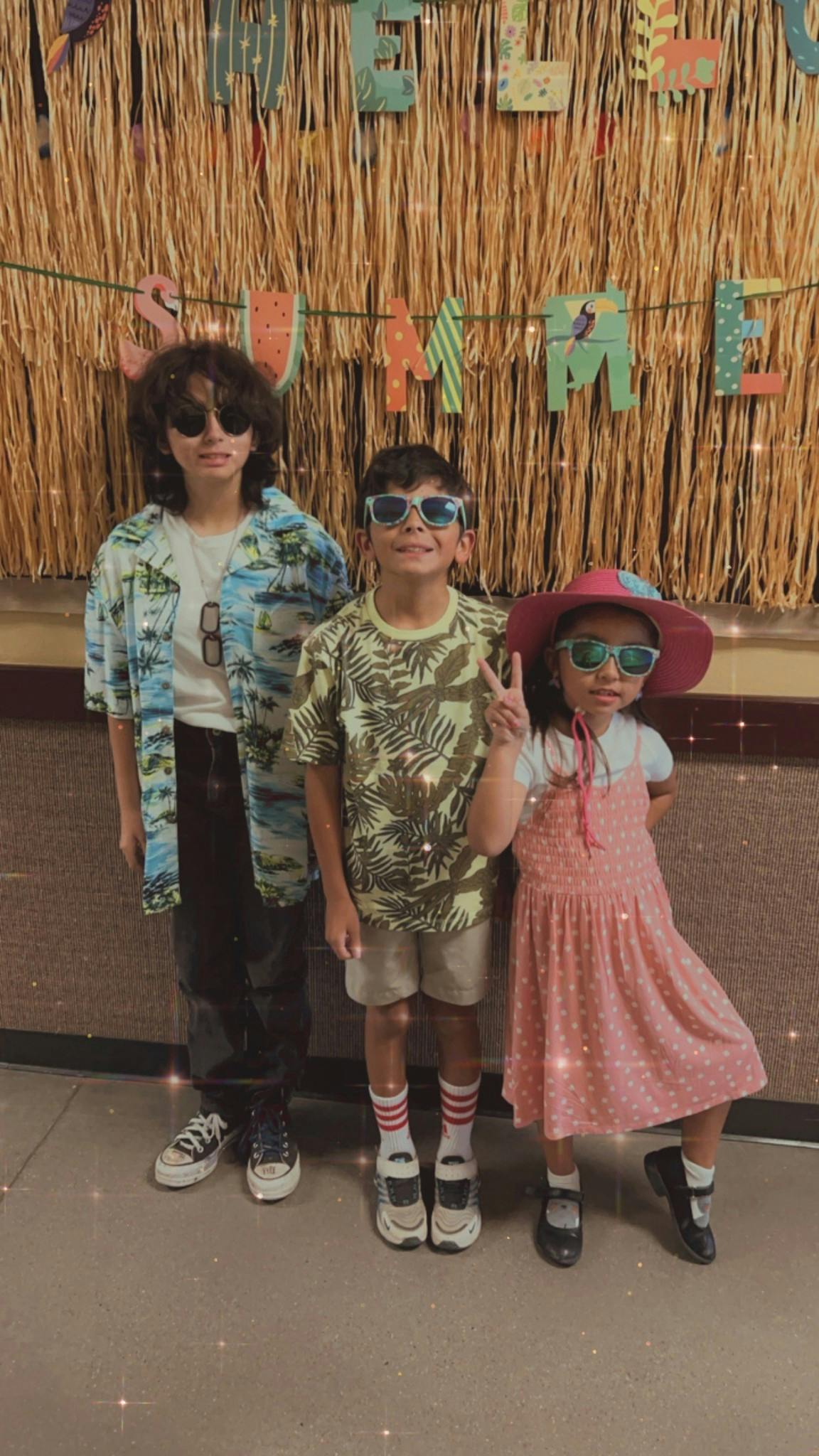 Three children wearing sunglasses and summer clothes in front of a straw wall