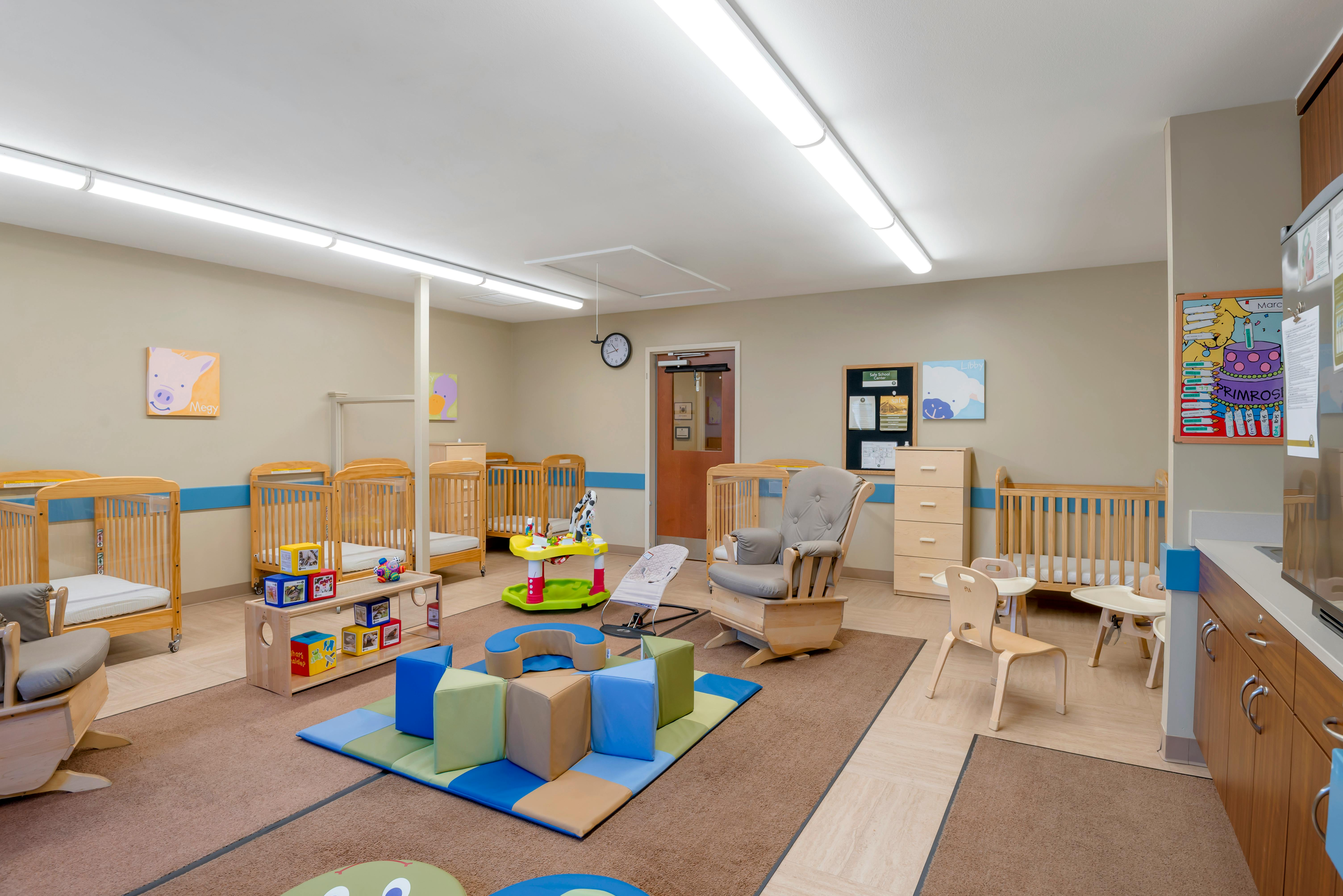 A brightly lit nursery with cribs, a play mat with soft blocks, and a rocking chair