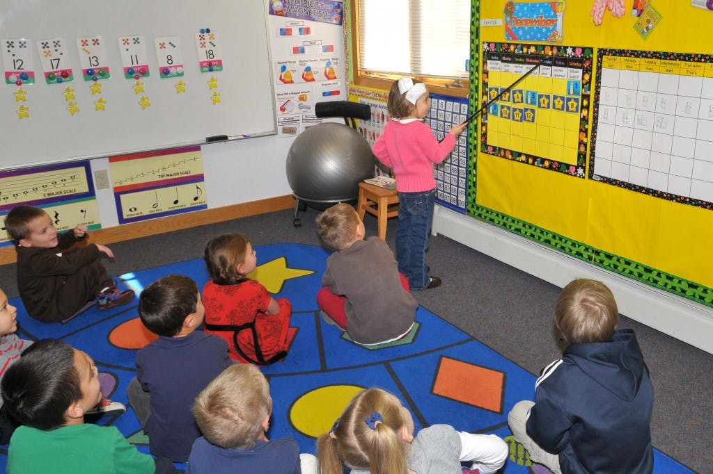 Child pointing at a board while other children sit on a rug in a classroom