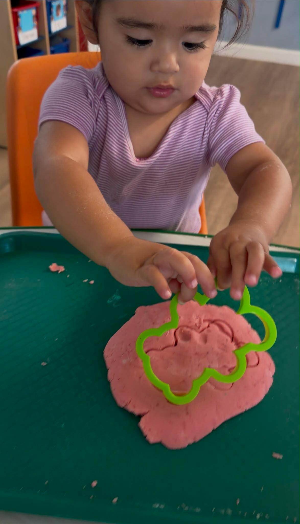 Child pressing a flower-shaped cutter into pink dough on a tray
