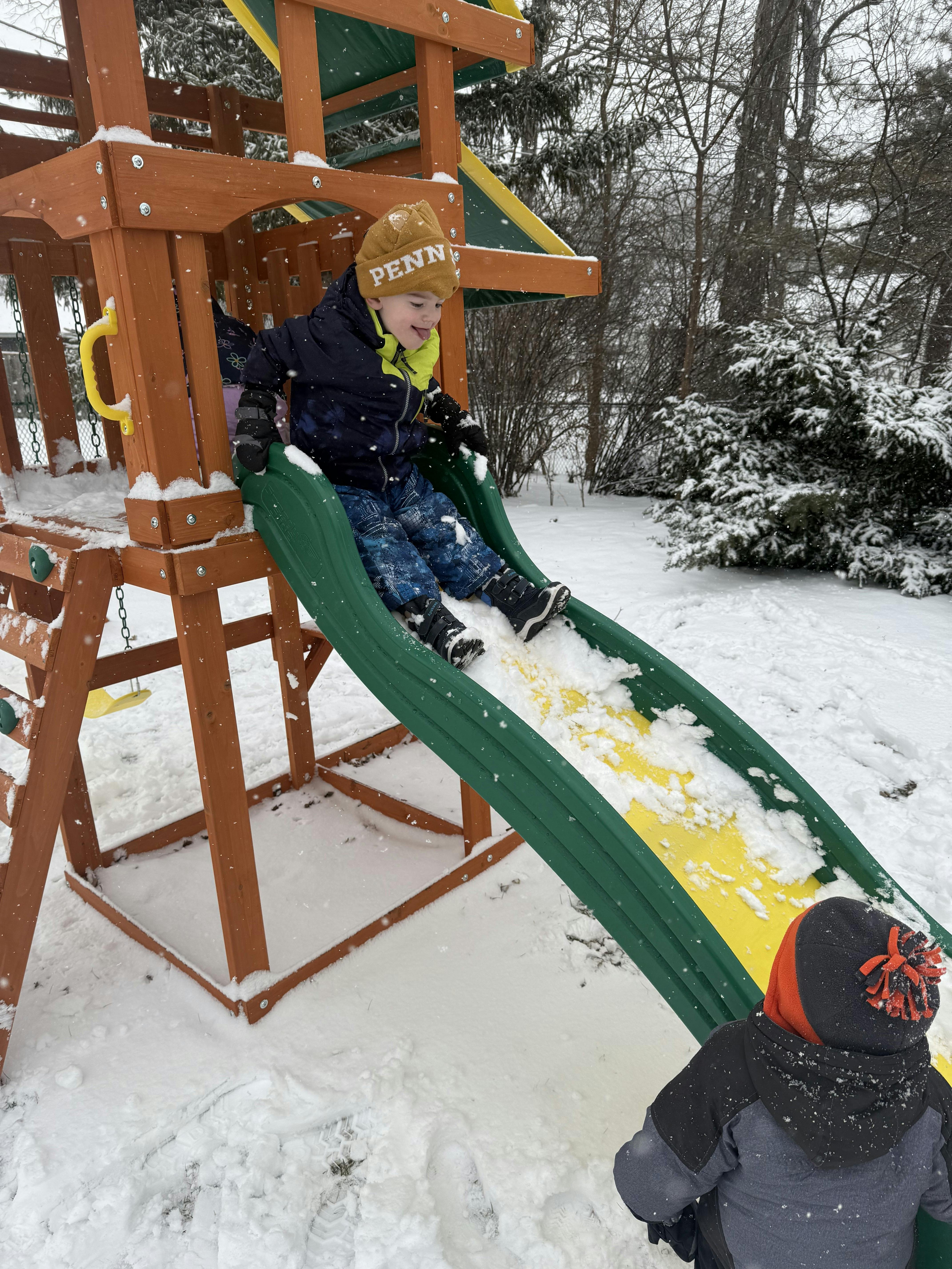 Child sliding down a snowy playground slide as another person watches