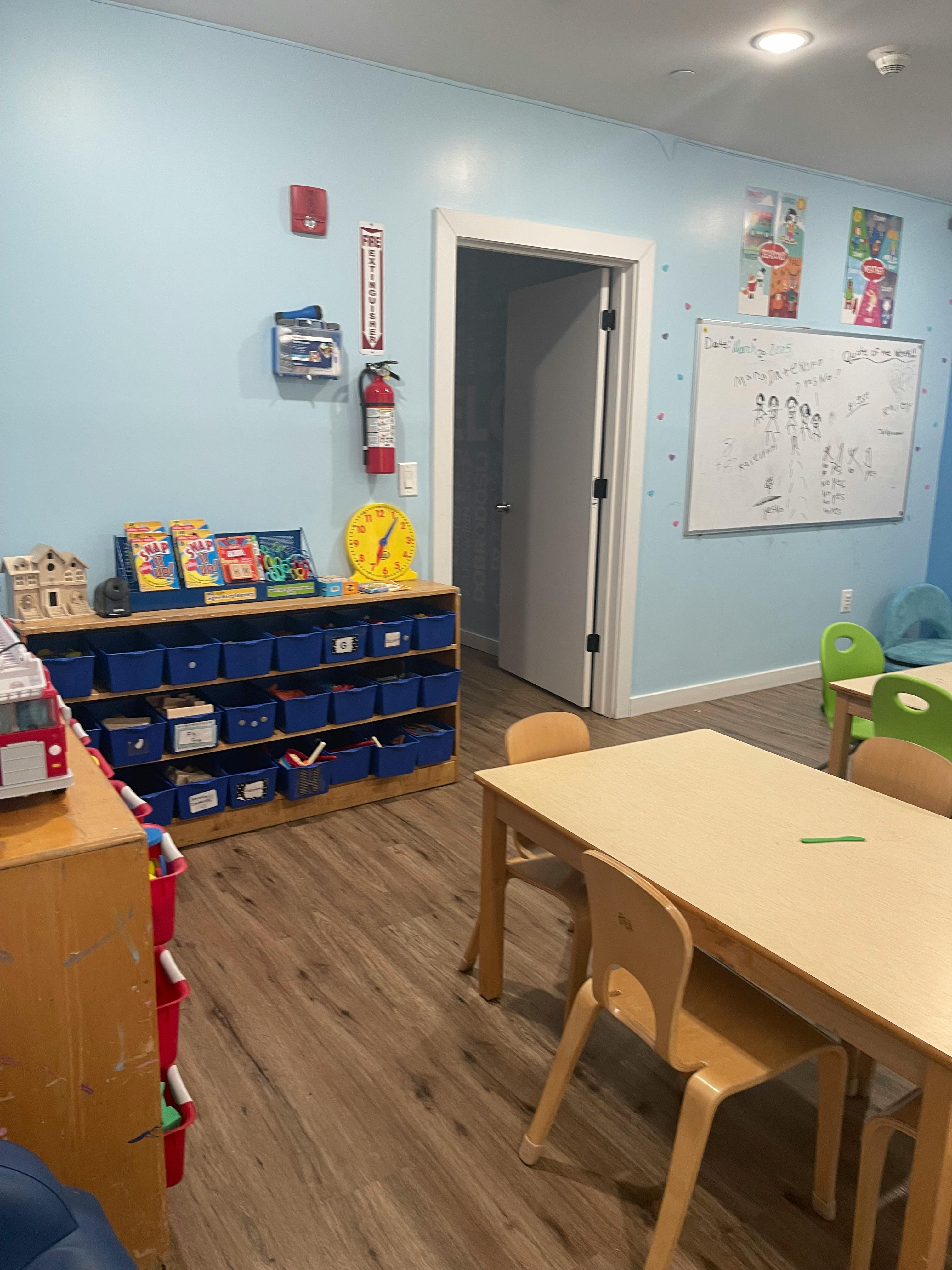 Classroom with small tables and chairs, storage unit with blue bins, and whiteboard