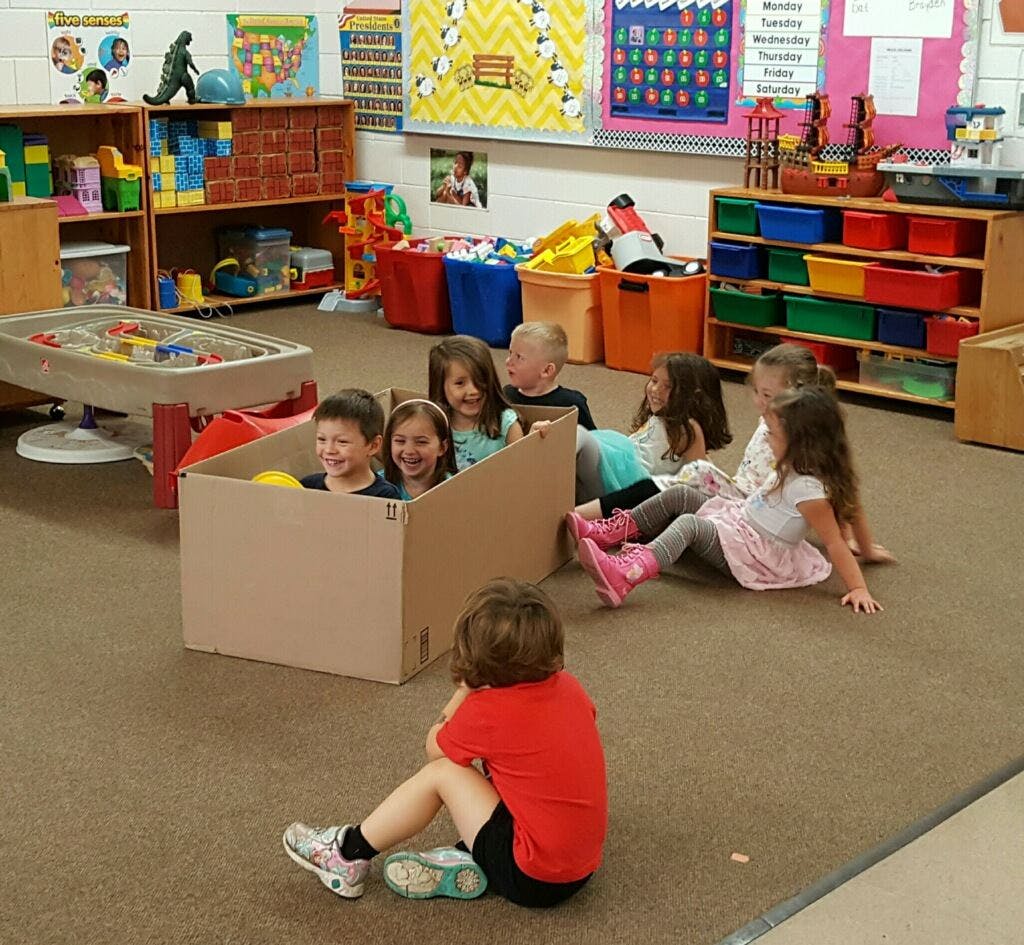 Children playing in a classroom, some in a cardboard box