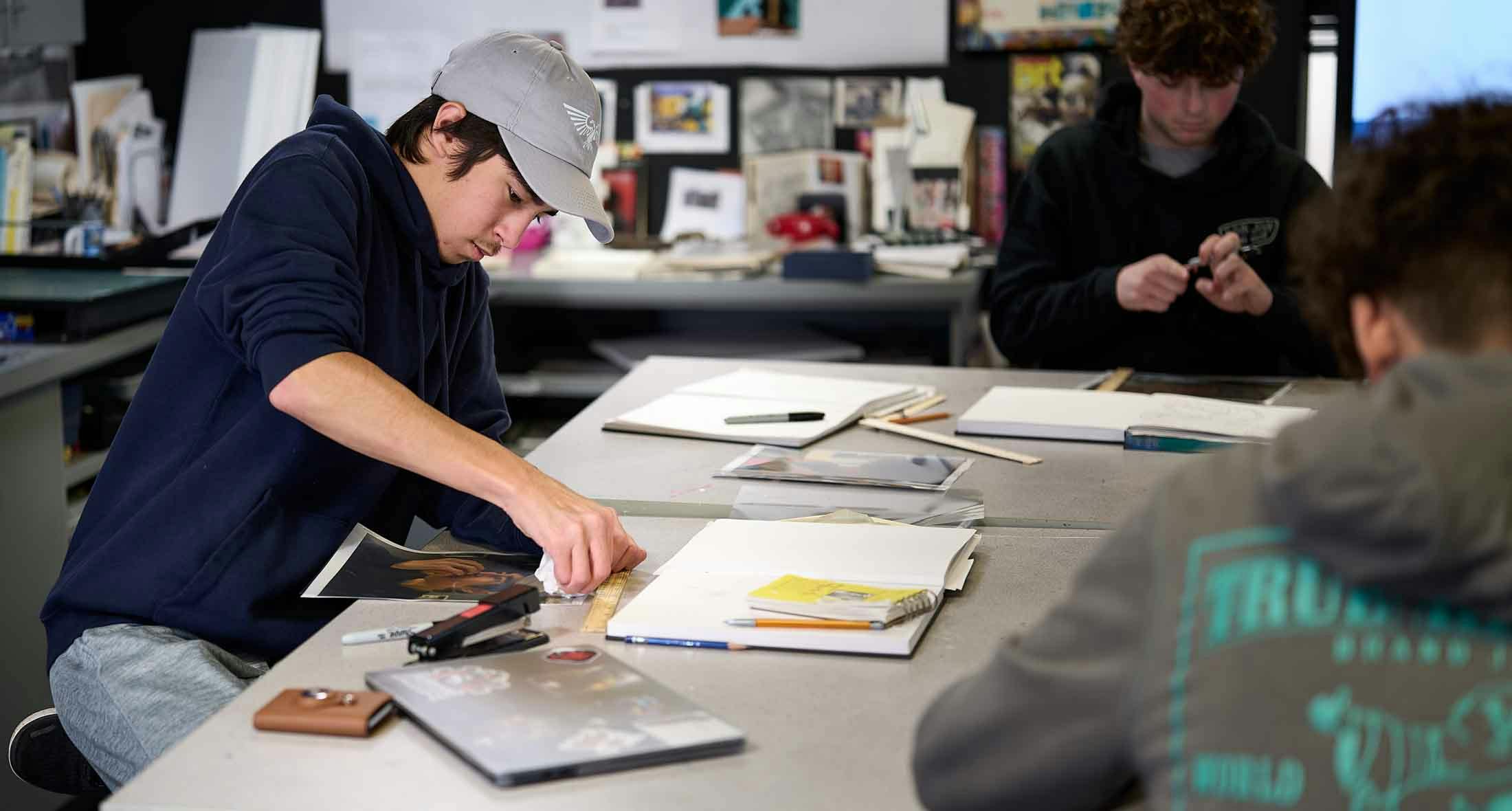 A person in a grey hat and navy hoodie working at a table in a classroom
