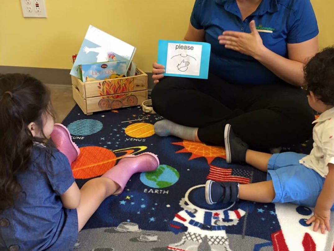 Person holding a card with "please" written on it, two children sitting on a rug