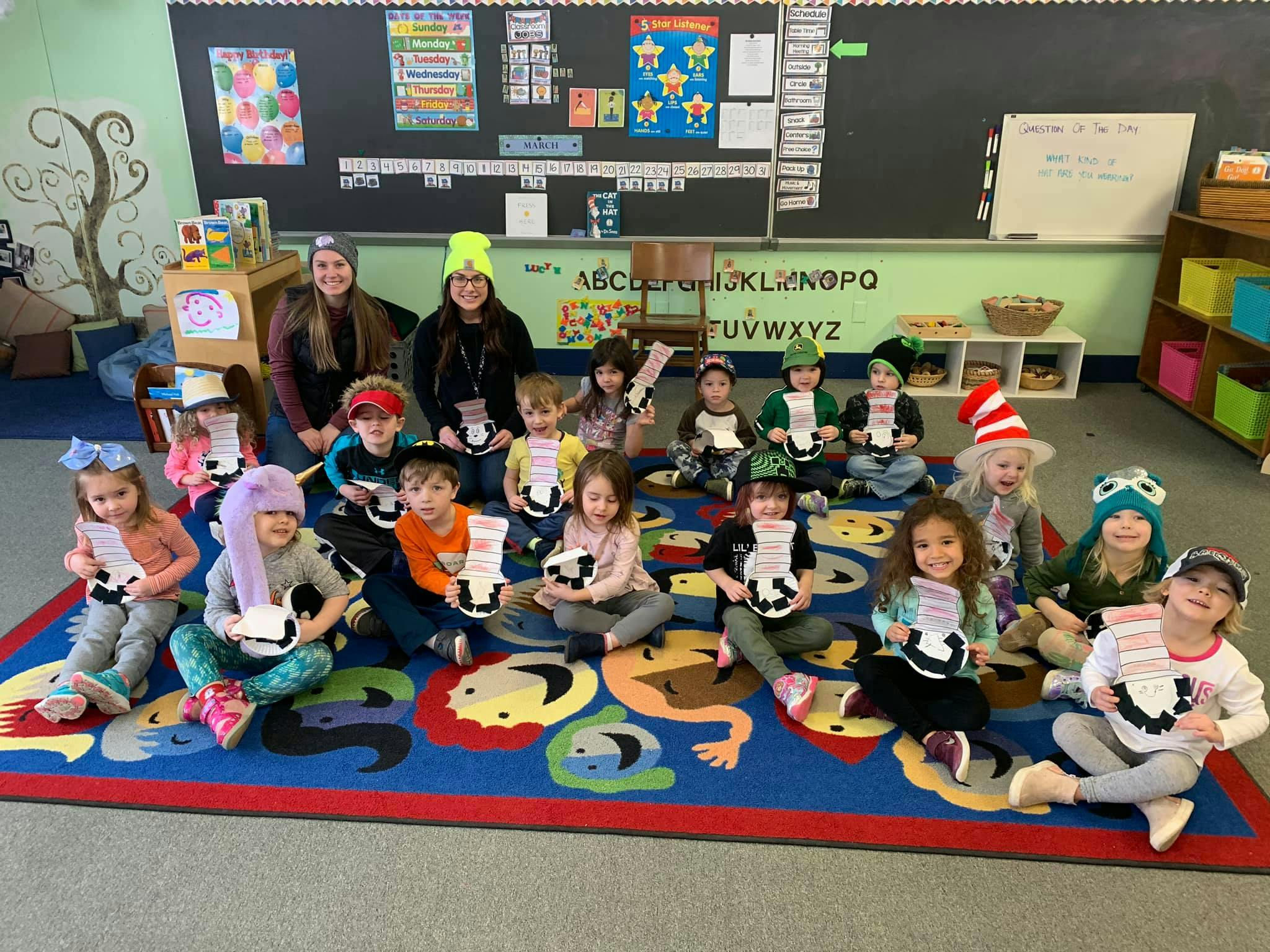 Two people and multiple children sitting on a colorful rug in a classroom