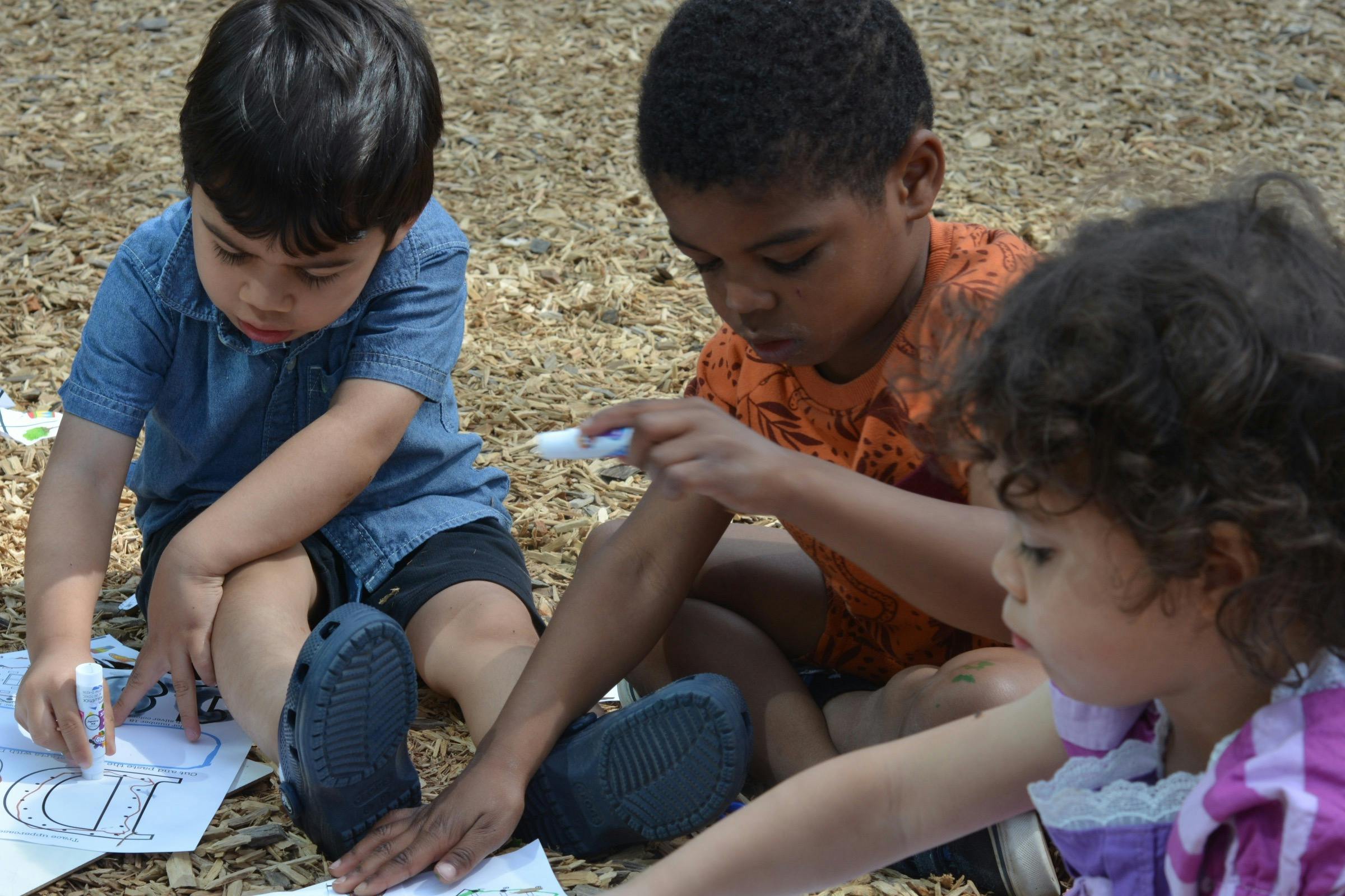 Three children sitting on wood chips and working on an activity with glue sticks