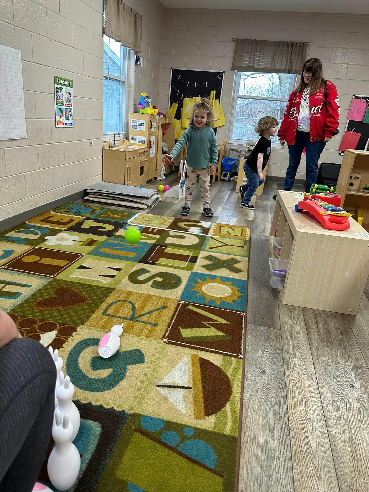 Child standing on an alphabet rug with a person and another child in the background