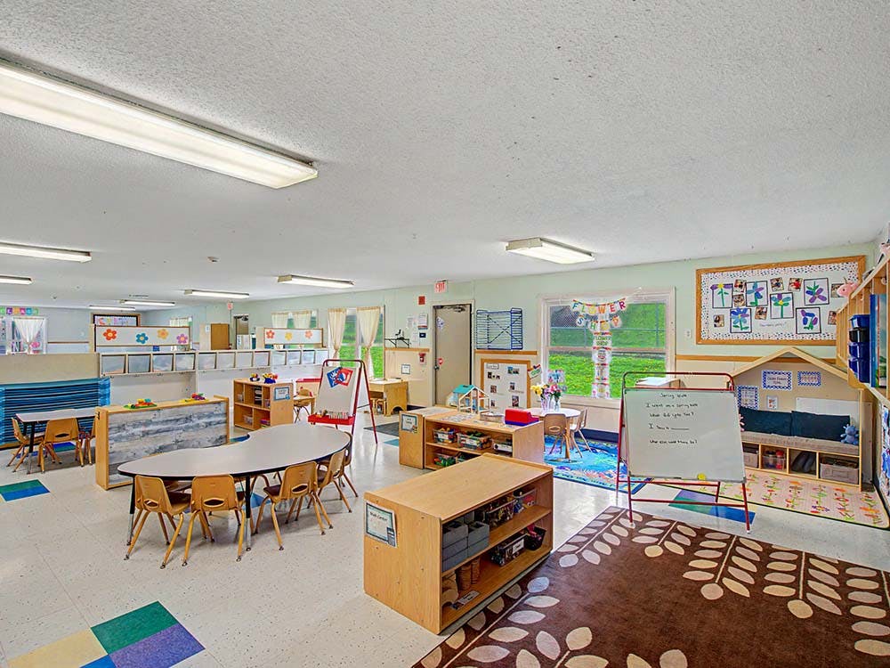 Empty preschool classroom with tables, chairs, shelving, and a whiteboard