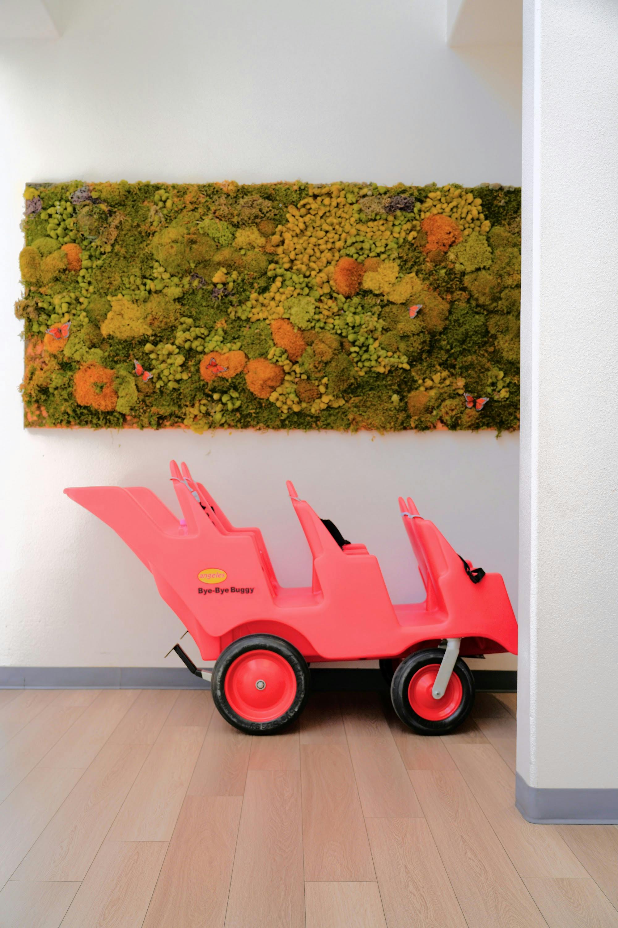A multi-seat Bye-Bye Buggy stroller parked in front of a moss wall art display