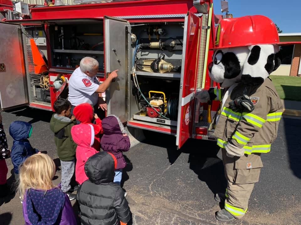 Firefighter showing equipment on a fire truck to children and a mascot