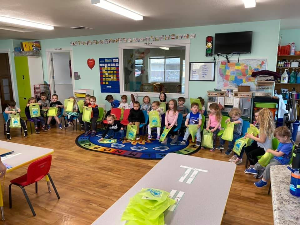 Children sitting on a rug in a classroom, holding green bags