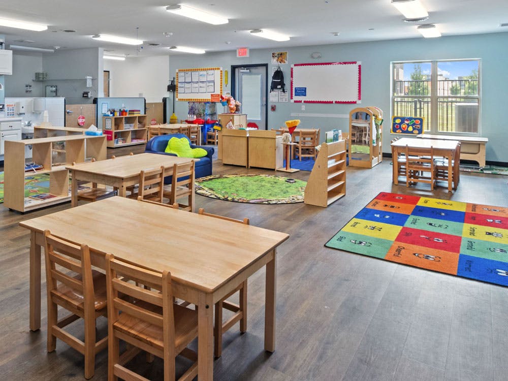 Empty classroom with small wooden tables, chairs, colorful rugs, and play areas